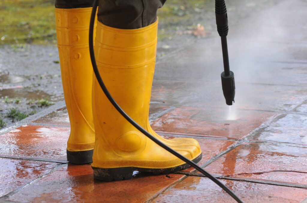 person wearing yellow rubber boots with high pressure water nozzle cleaning dirt tiles Pool Deck & Enclosure Cleaning Services in Tampa FL